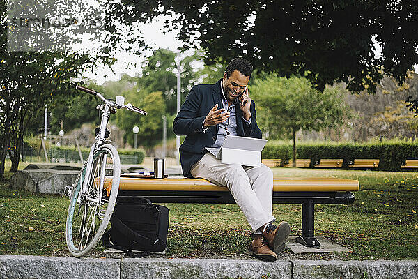 Männlicher Freiberufler in voller Länge  der im Park sitzt und mit seinem Smartphone spricht