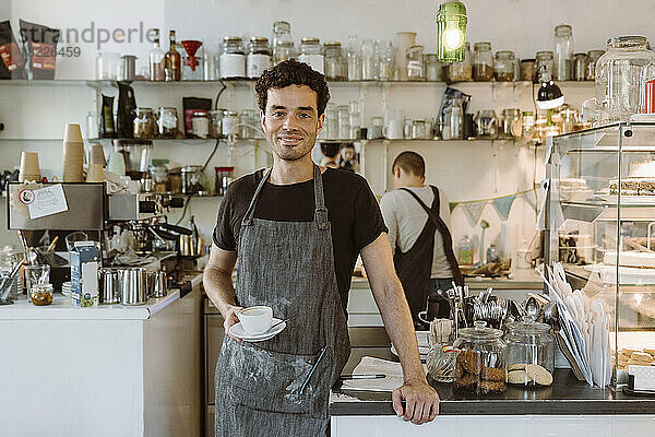 Lächelnder männlicher Besitzer mit Schürze  der eine Kaffeetasse in einem Café hält