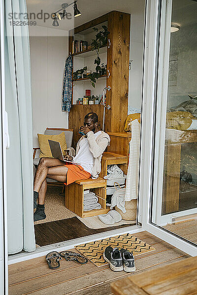 Mann mit Laptop trinkt Kaffee und sitzt auf einem Holzstuhl zu Hause