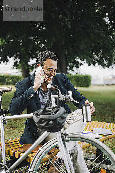 Glücklicher Geschäftsmann  der mit dem Handy telefoniert  während er mit dem Fahrrad im Park sitzt