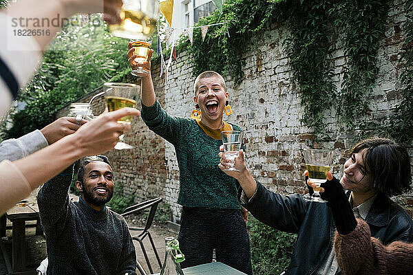 Fröhliche Frau mit offenem Mund stößt mit Freunden während einer Dinnerparty im Hinterhof auf Getränke an