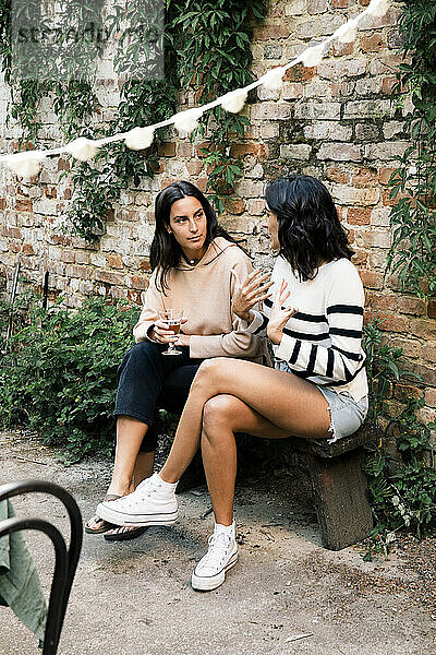 Weibliche Freunde unterhalten sich auf einer Bank in der Nähe einer Backsteinmauer bei einer Gartenparty
