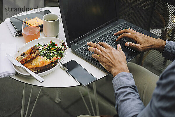 Hände eines Geschäftsmannes  der beim Essen einen Laptop benutzt und ein Smartphone auf dem Tisch liegen hat