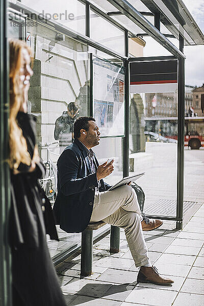 Männlicher Profi in voller Länge mit Laptop an der Bushaltestelle an einem sonnigen Tag sitzend
