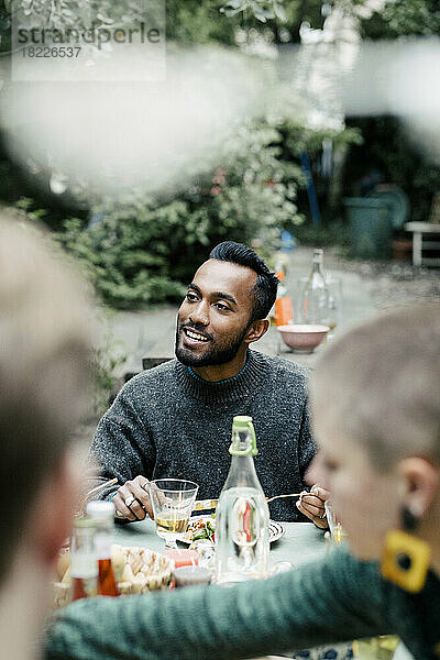 Lächelnder Mann  der am Esstisch während einer Dinnerparty im Hinterhof sitzt und nachdenkt