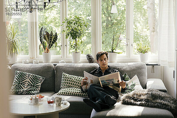 Junger Mann liest Zeitung  während er zu Hause auf dem Sofa im Wohnzimmer sitzt