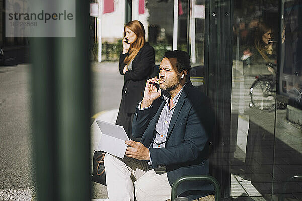 Männlicher Unternehmer  der an einem sonnigen Tag an einer Bushaltestelle mit seinem Laptop sitzt und mit seinem Smartphone spricht