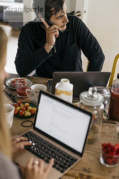 Männlicher Freiberufler  der mit seinem Smartphone telefoniert  während er zu Hause auf dem Tisch sitzt und frühstückt