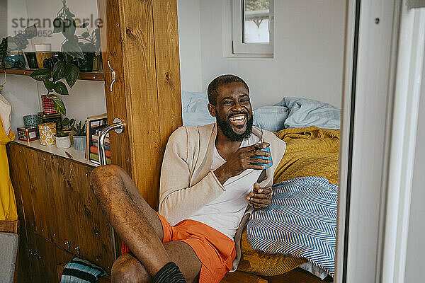 Glücklicher reifer Mann mit Kaffeetasse sitzt mit gekreuzten Beinen zu Hause