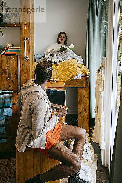 Seitenansicht eines Mannes  der sich mit einer Frau unterhält  die ein Buch zu Hause auf dem Bett liegen hat
