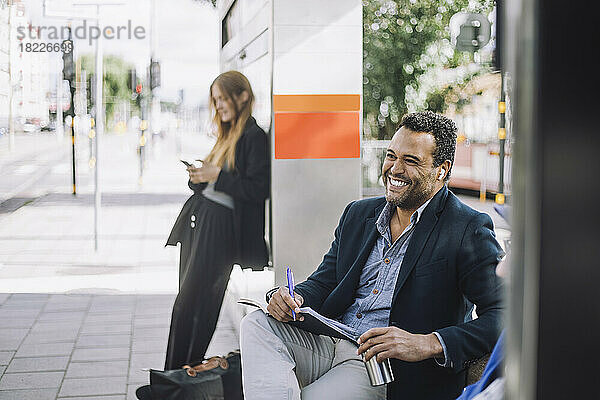 Glücklicher männlicher Freiberufler mit kabellosen In-Ear-Kopfhörern  der mit einem Tagebuch gegen eine Geschäftsfrau an der Bushaltestelle sitzt