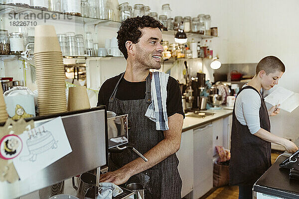 Lächelnder Besitzer mit Schürze  der wegschaut  während er in der Nähe der Kaffeemaschine im Café steht