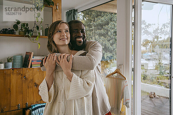 Porträt eines lächelnden Mannes  der seine Freundin umarmt  während sie zusammen zu Hause stehen