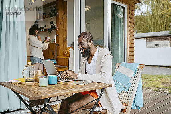 Glücklicher reifer Mann  der einen Laptop benutzt  während er am Tisch auf der Veranda vor dem Haus sitzt