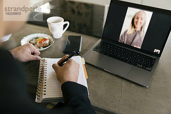 Hände eines Geschäftsmannes  der in ein Tagebuch schreibt  während er einen Videoanruf mit einer Kollegin auf einem Laptop führt
