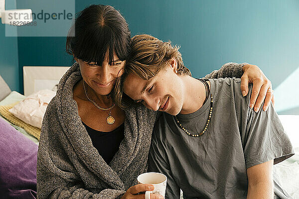 Lächelnde Mutter mit Arm um den Sohn  der sich auf die Schulter stützt  auf dem Bett zu Hause sitzend