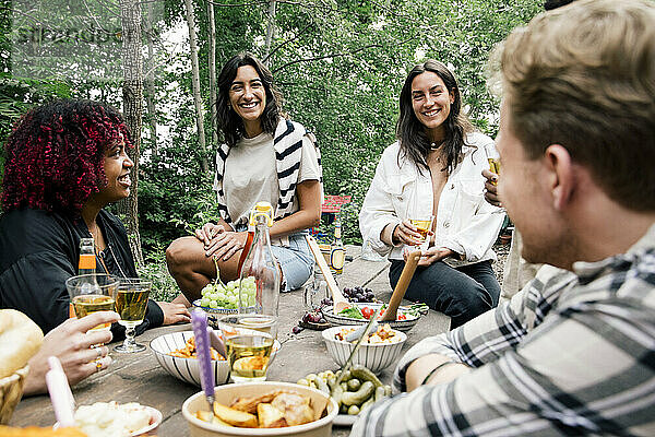 Lächelnde Frauen im Gespräch mit Freunden mit Essen auf dem Tisch während einer Gartenparty im Hinterhof
