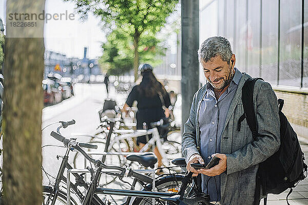 Lächelnder reifer Geschäftsmann  der ein Smartphone benutzt  während er an einer Fahrradabstellanlage steht