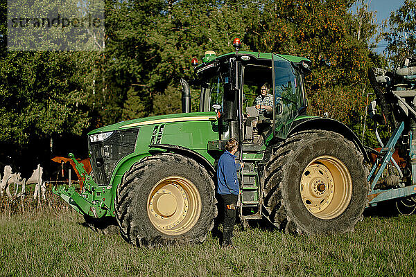 Landwirt sitzt im Traktor und diskutiert mit einem Mann  der an einem sonnigen Tag im Feld steht