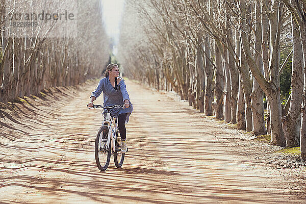 Teenager-Mädchen (16–17) fährt Fahrrad auf einer von Bäumen gesäumten Straße