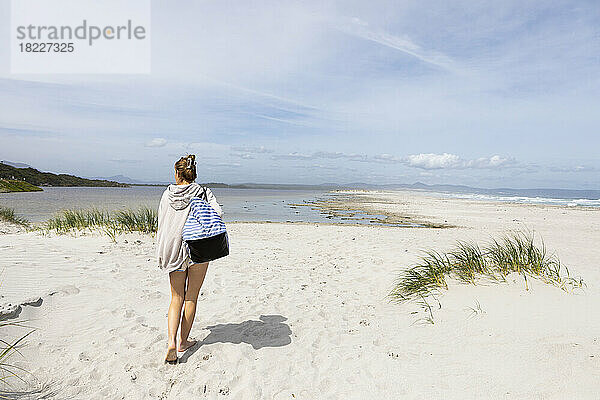 Südafrika  Hermanus  Teenager-Mädchen (16-17) mit Strandtasche spaziert am Sandstrand
