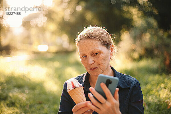 Reife Frau mit Smartphone und Eis steht im Park
