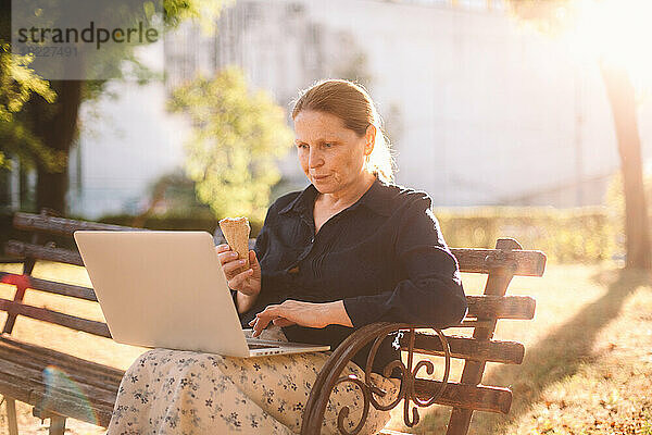 Reife Geschäftsfrau isst Eis mit Laptop-Computer im Park