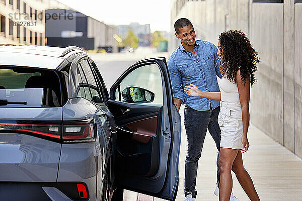 Lächelnder Mann öffnet Tür eines Elektroautos für Freundin am Straßenrand