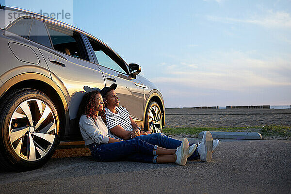 Lächelnde Frau sitzt bei Sonnenuntergang mit ihrem Freund vor dem Auto