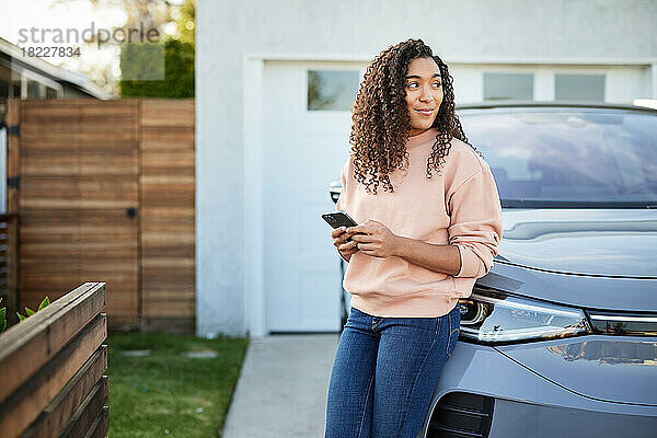 Contemplative woman with smart phone leaning on electric car