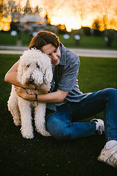 Teenager umarmt goldenen Doodle-Hund vor Sonnenuntergangsbäumen