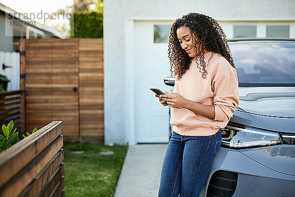 Smiling woman using smart phone leaning on electric car in front yard