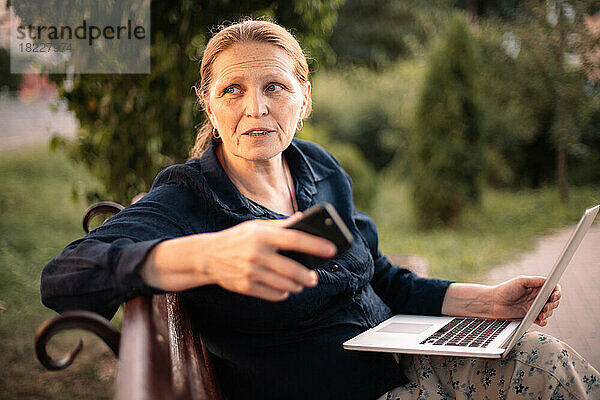 Reife Geschäftsfrau mit Laptop und Smartphone sitzt im Park