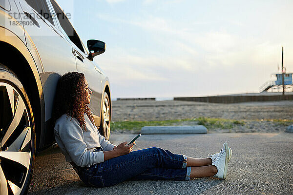 Junge Frau mit Smartphone stützt sich auf Elektroauto auf Parkplatz