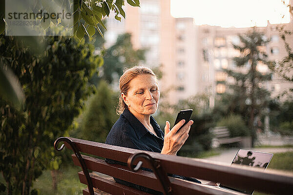 Glückliche reife Geschäftsfrau nutzt Smartphone und Laptop im Park