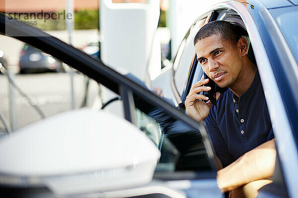 Mittlerer erwachsener Mann  der im Elektroauto sitzt und auf einem Smartphone spricht