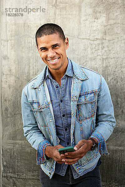 Porträt eines lächelnden Mannes mit Smartphone  der an der Wand steht