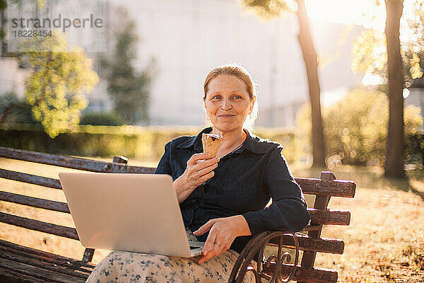 Glückliche ältere Geschäftsfrau mit Laptop und Eis im Park