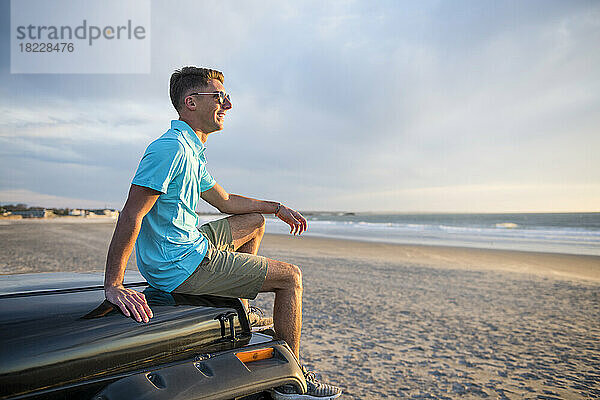 Junger Mann mit Sonnenbrille auf der Motorhaube des Autos bei Sonnenaufgang am Strand