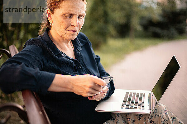 Reife Geschäftsfrau benutzt Smartphone und Laptop im Park