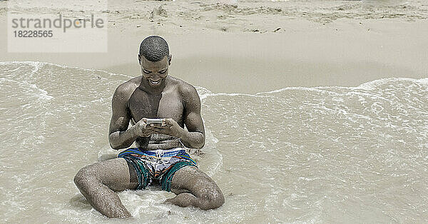 Afroamerikanischer Mann benutzt ein Smartphone in der Nähe der Meereswellen
