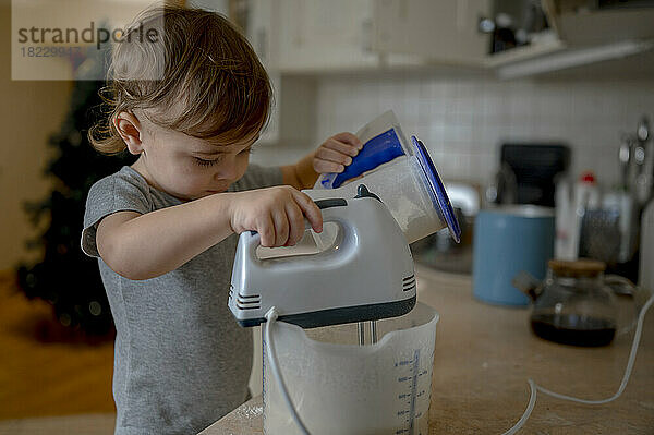 Ein kleiner Junge gießt Mehl in einen Behälter und mischt zu Hause mit einem Elektromixer