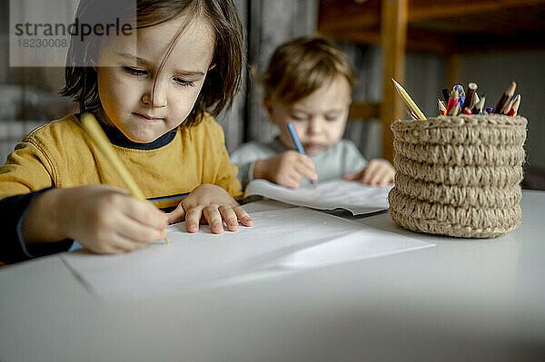 Junge mit Bruder zeichnet mit Buntstiften in einem Jutebehälter auf Papier