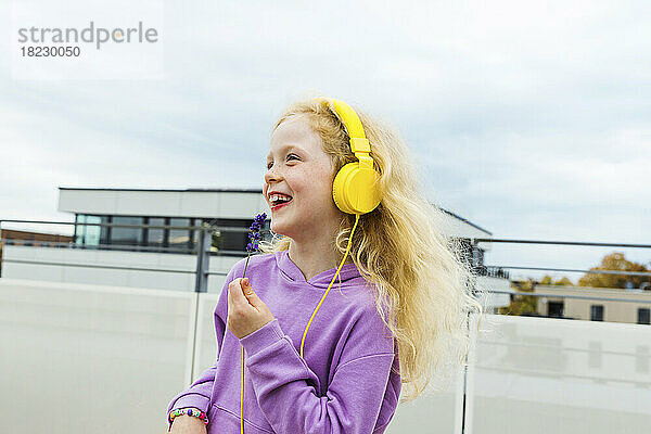 Fröhliches Mädchen hält eine kleine Blume in der Hand und hört Musik über Kopfhörer auf dem Dach