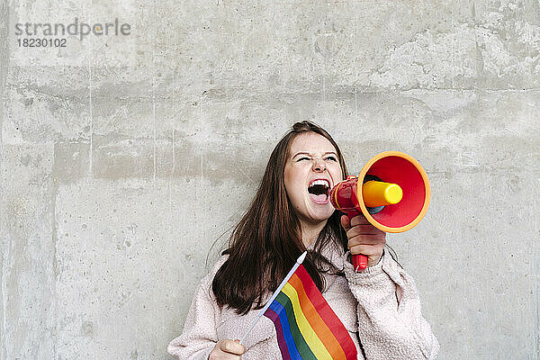 Junge Frau hält Regenbogenfahne in der Hand und schreit durch Megafon  das an die Wand lehnt