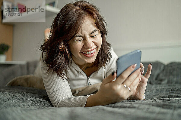 Fröhliche junge Frau mit Smartphone liegt zu Hause im Bett