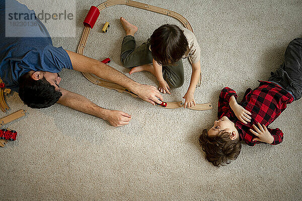 Junge schaut Vater und Bruder an  die mit einer hölzernen Spielzeugeisenbahn auf dem Boden spielen