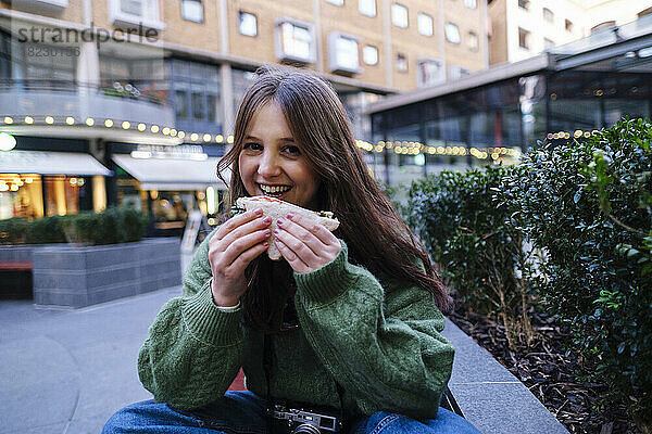Glückliche junge Frau isst Sandwich vor Gebäuden