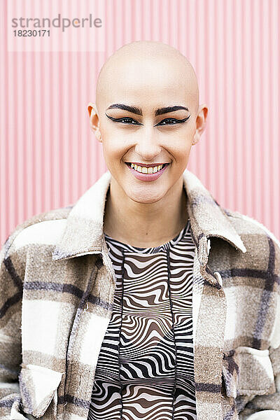 Fröhliche junge Frau mit rasiertem Kopf vor rosa Wand