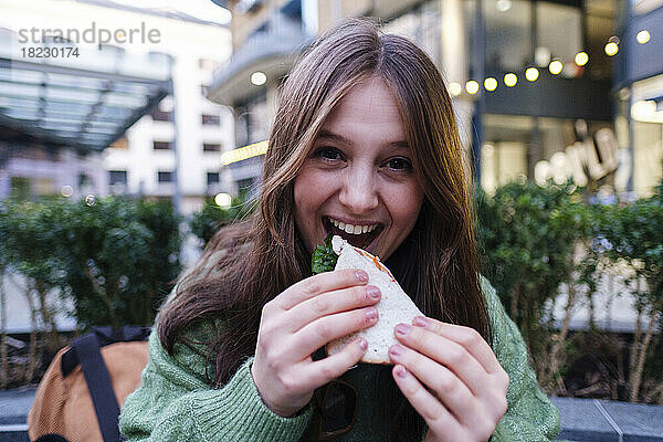 Glückliche junge Frau isst Sandwich vor dem Gebäude